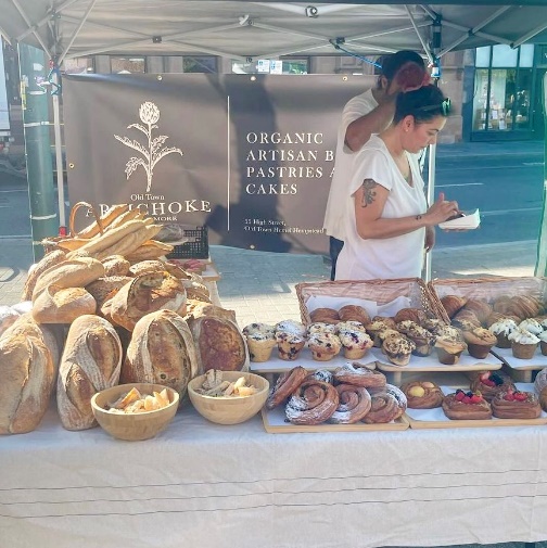 Artisan bakery stall at St Albans Market displaying loaves, pastries and cakes, with traders serving customers beneath a canopy.